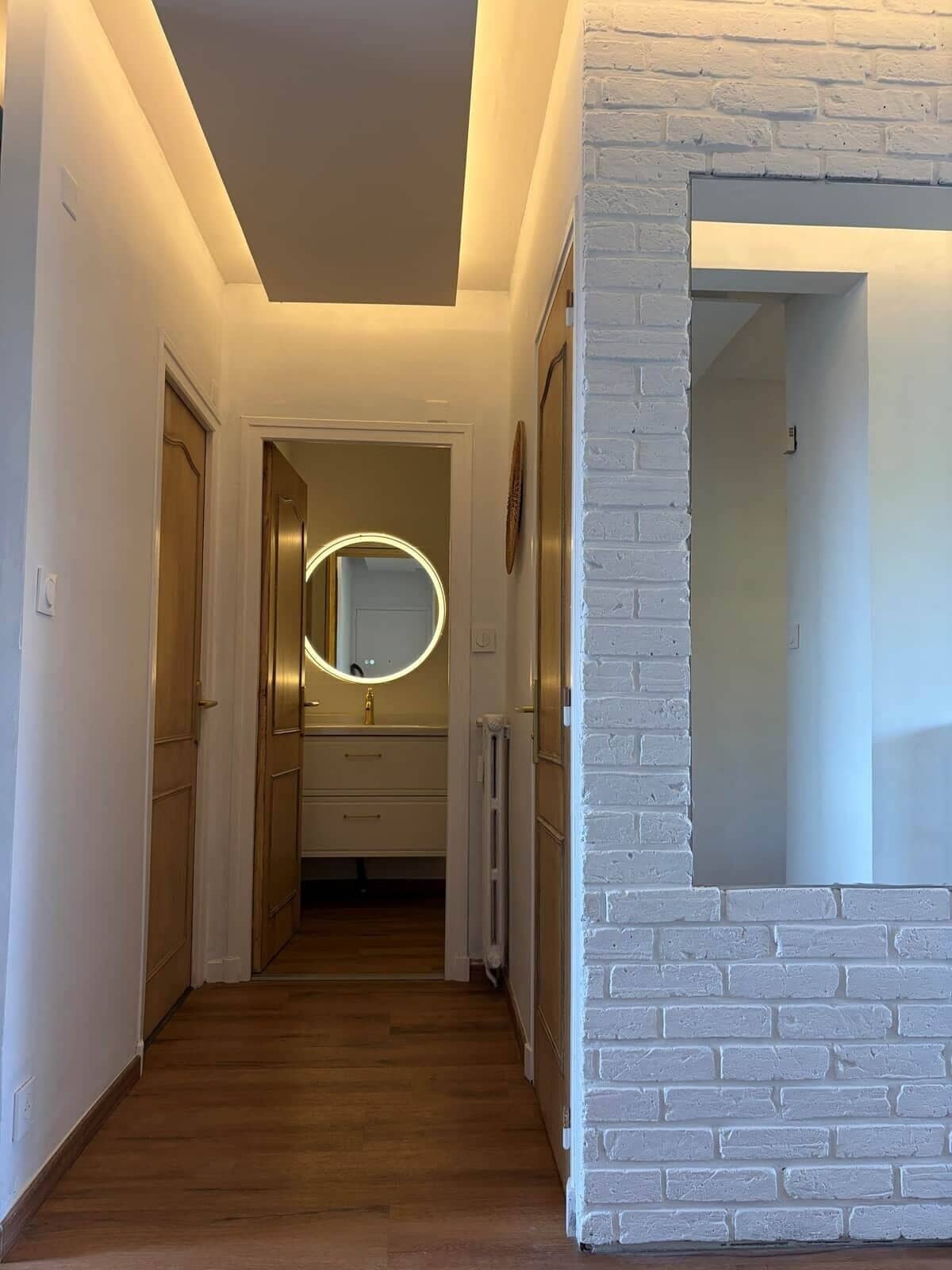 A hallway with wooden flooring, a white brick accent wall, and a round mirror with lighting.