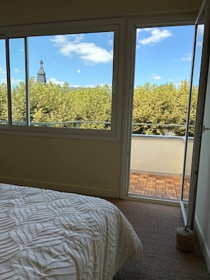 A bedroom with a large window, a bed, and a view of a building and trees. - Le Balcon de Sarlat * Private parking & Town center 25 meters away (Sarlat-la-Canéda)
