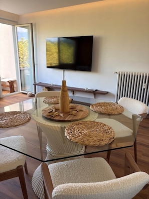 A modern living room with a glass table surrounded by chairs, a flat-screen TV mounted on the wall, and a radiator. - Le Balcon de Sarlat * Private parking & Town center 25 meters away (Sarlat-la-Canéda)
