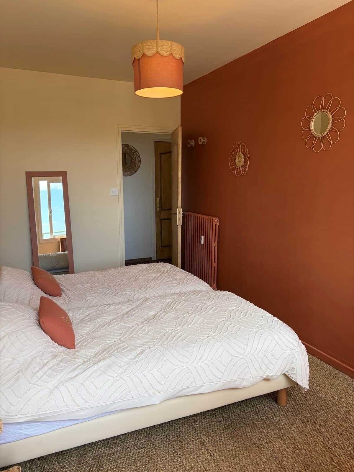 Une chambre avec un grand lit, un luminaire suspendu et une porte donnant sur une autre pièce.