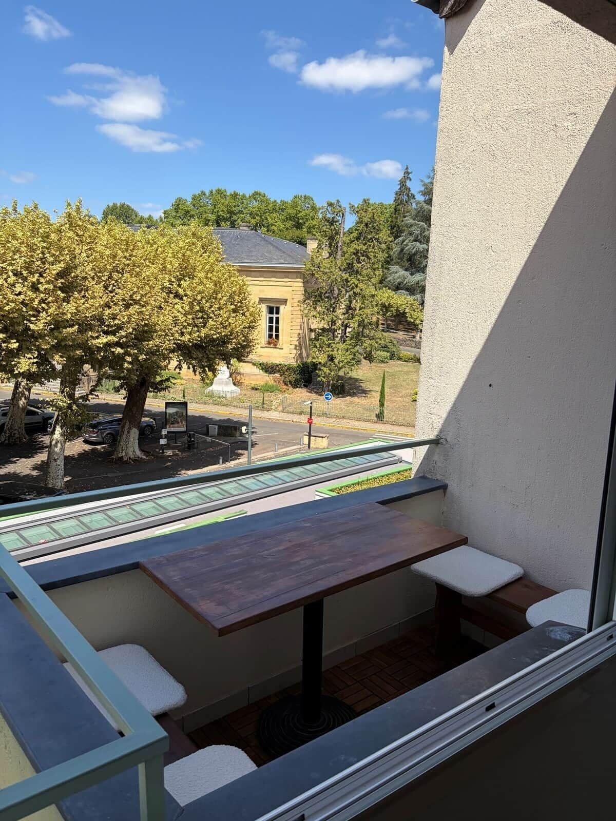 A balcony with a table and seating, overlooking a courtyard with trees and a building.