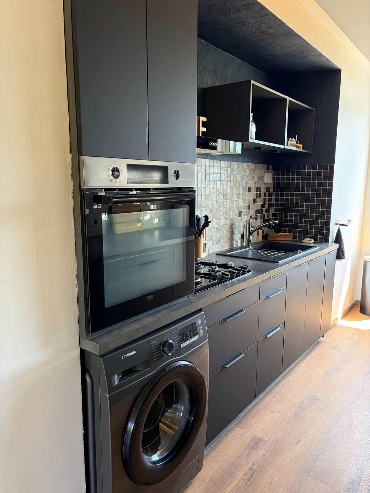 A compact kitchen with a built-in oven, microwave, and washing machine, featuring dark cabinetry and a tiled backsplash.
