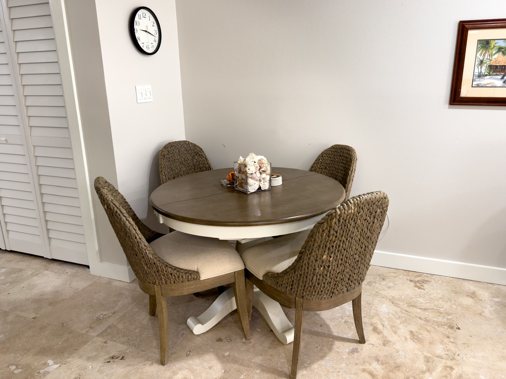 Une table de salle à manger ronde, entourée de quatre chaises en osier, une horloge au mur et un tableau encadré.