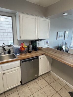 A kitchen with white cabinets, a dishwasher, a wooden countertop, and a window with blinds.