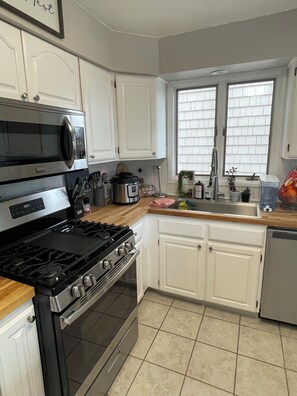 A modern kitchen with white cabinets, stainless steel appliances, a window with blinds, and a tiled floor.