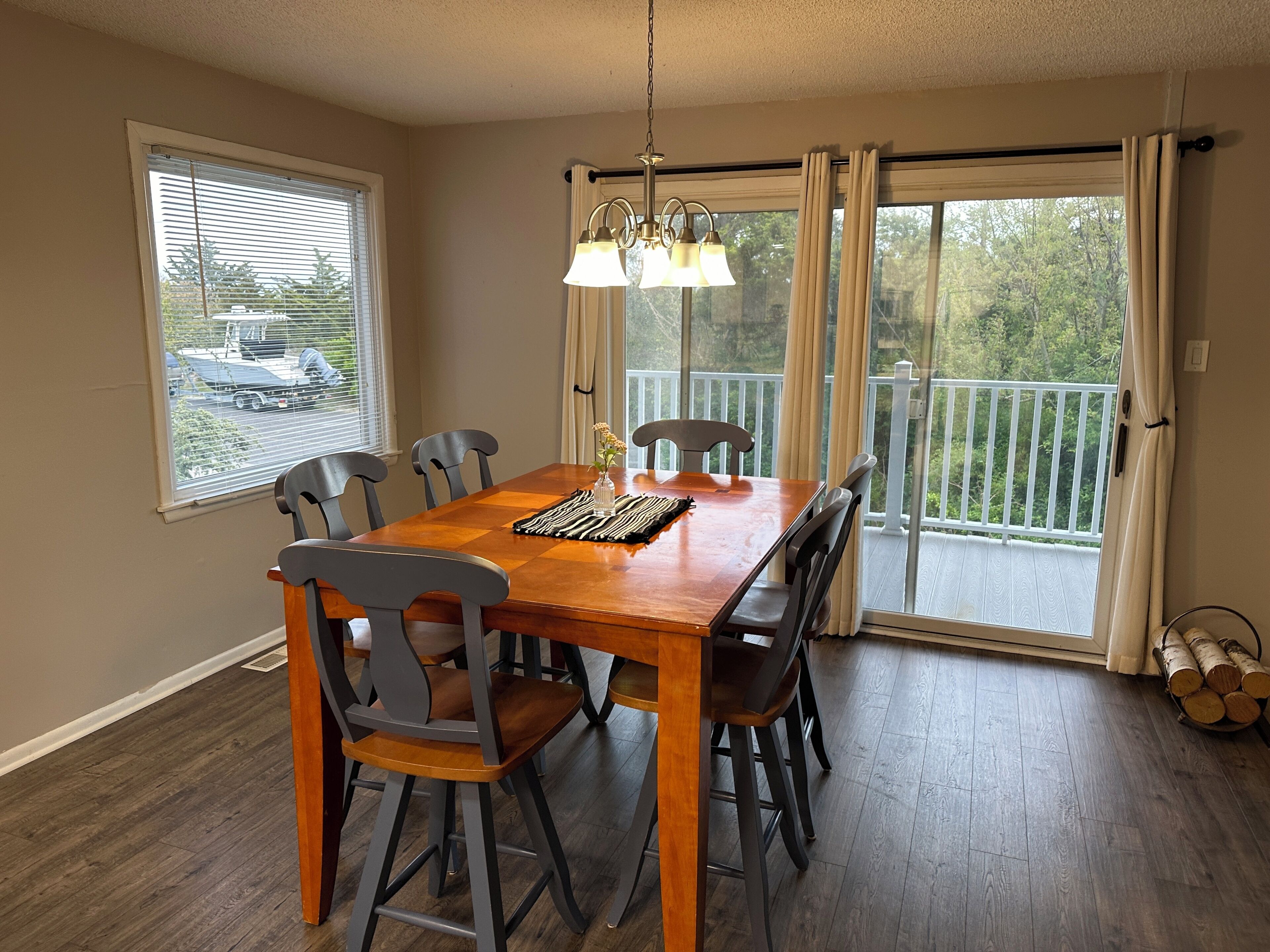 A dining room with a wooden table, six chairs, a chandelier, and a view of a balcony with a railing.