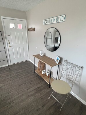 A room with a white door, a round mirror, a small table with a picture frame, and a metal chair.