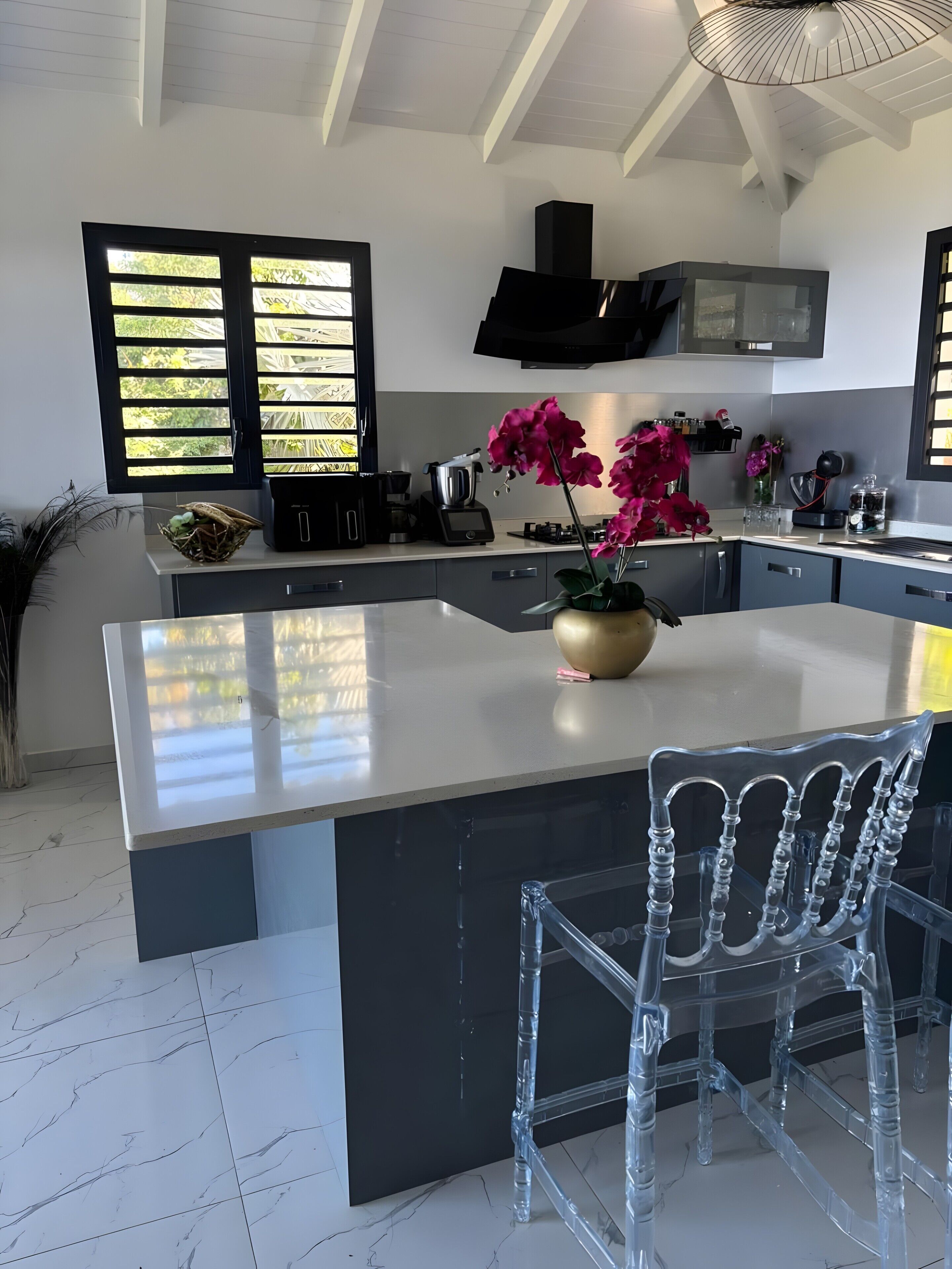 A modern kitchen with a large island, black cabinetry, and a clear chair.