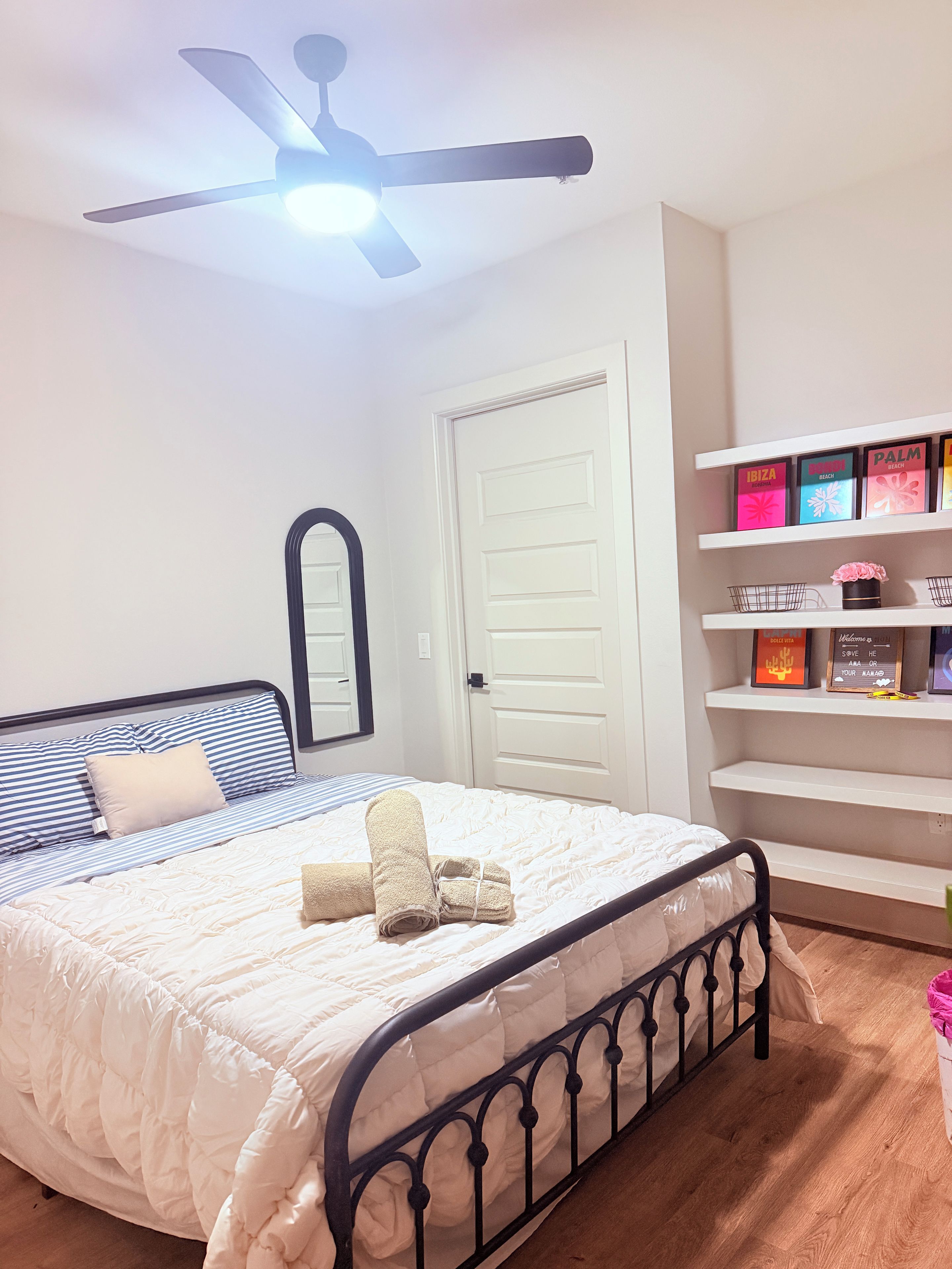 A bedroom with a bed, a ceiling fan, a door, and a shelf with books.