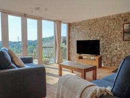 Una sala de estar con pared de piedra, un televisor de pantalla plana sobre un pedestal de madera y un amplio ventanal corredizo que da a una terraza.