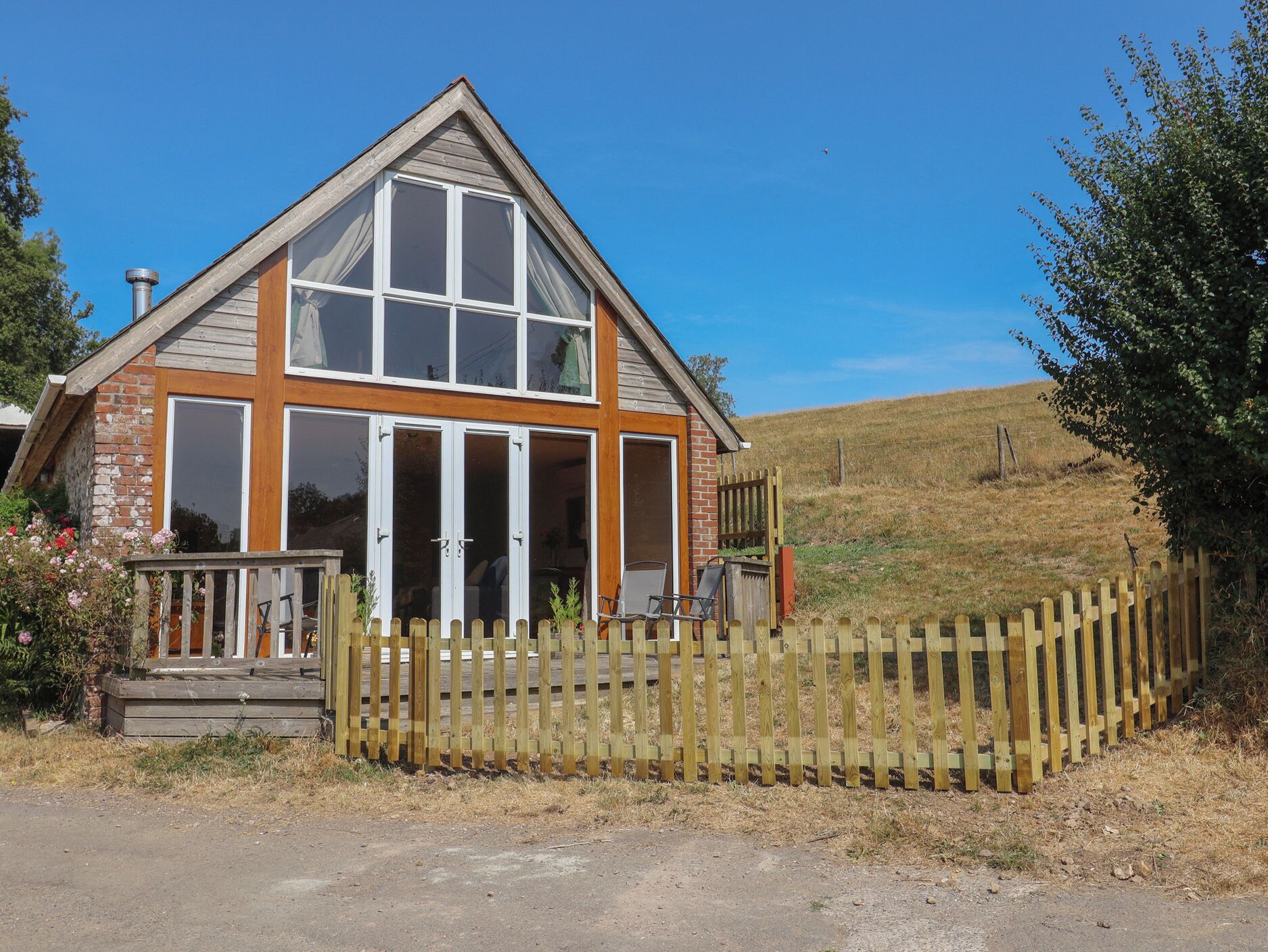 Ein zweistöckiges Haus mit einer Holzterrasse, großen Fenstern und einem eingezäunten Garten.