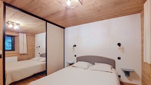 Un dormitorio con un espejo grande, una cama con ropa de cama blanca, dos mesitas de noche con lámparas y una ventana con cortinas.