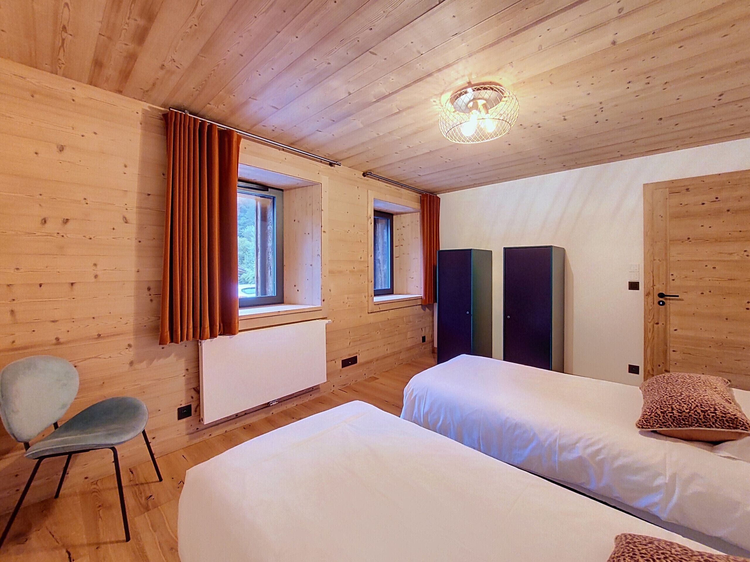 Quarto com duas camas, uma cadeira, um radiador e uma janela com cortinas.