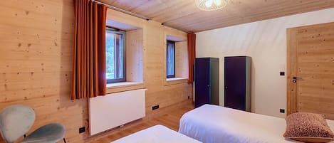 Quarto com duas camas, uma cadeira, um radiador e uma janela com cortinas.