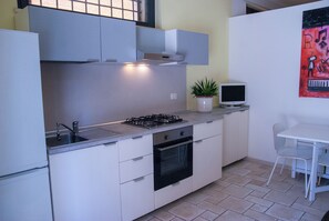 A compact kitchen with a gas stove, oven, and a small dining area with a white table and chair. - "Le casette del Papero" Vacation Home: Relax Among the Hills and the Sea (Montemarciano)