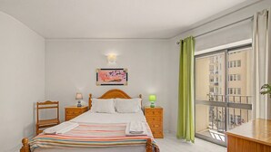 Quarto de hotel com cama, duas mesas de cabeceira, um criado-mudo, janela com vista para edifícios e luminária fixada na parede.