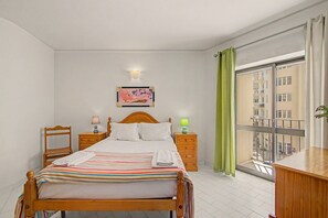 A hotel room with a bed, two bedside tables, a nightstand, a window with a view of buildings, and a wall-mounted light fixture. - Apartment 'Gabriel 1' with Shared Pool and Balcony (Armação de Pêra)