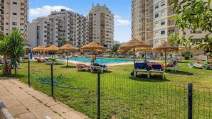 Área de piscina com espreguiçadeiras e guarda-sóis, cercada por edifícios altos.