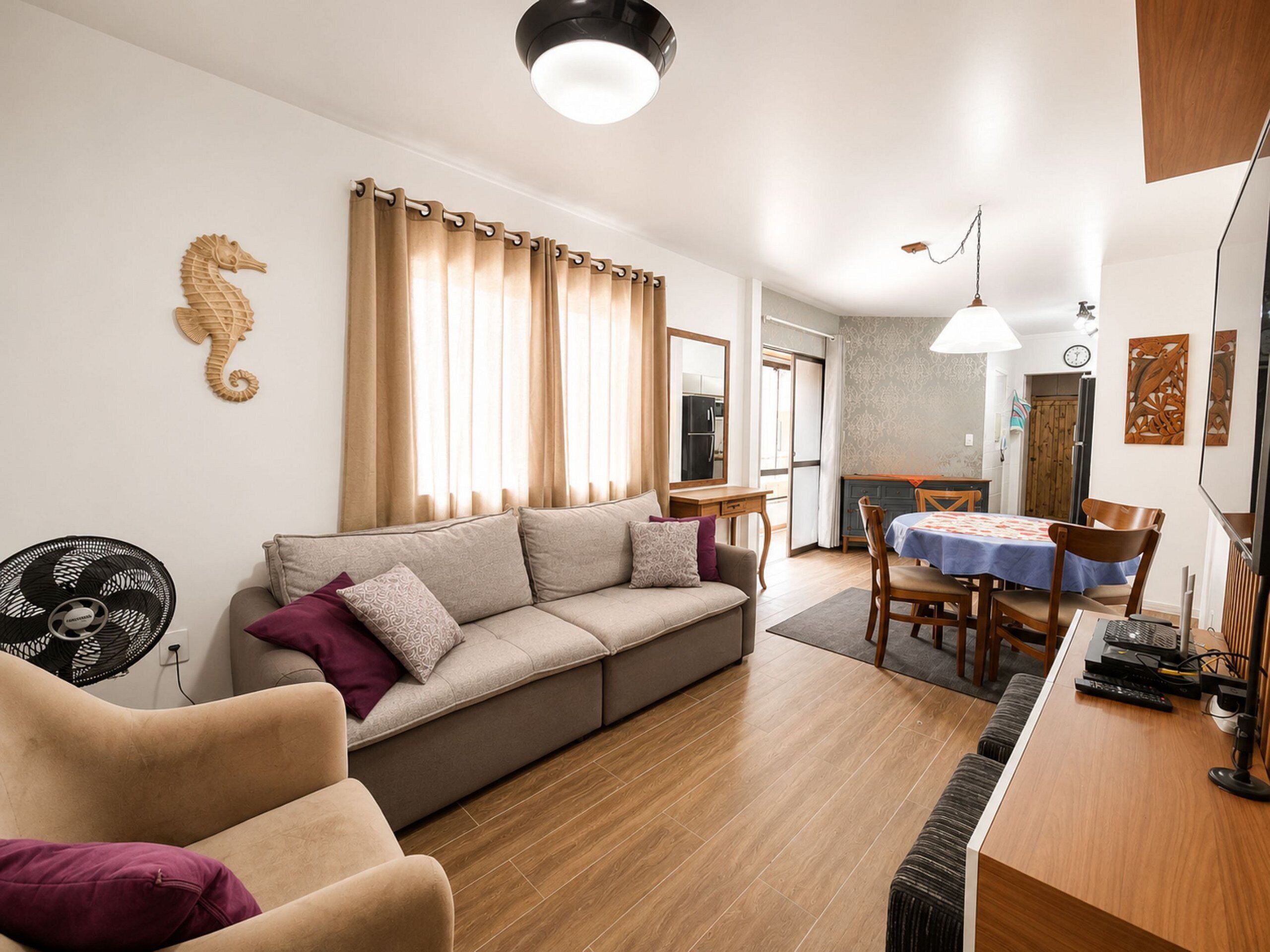 A living room with a sofa, a dining table with chairs, a fan, and a wooden floor.