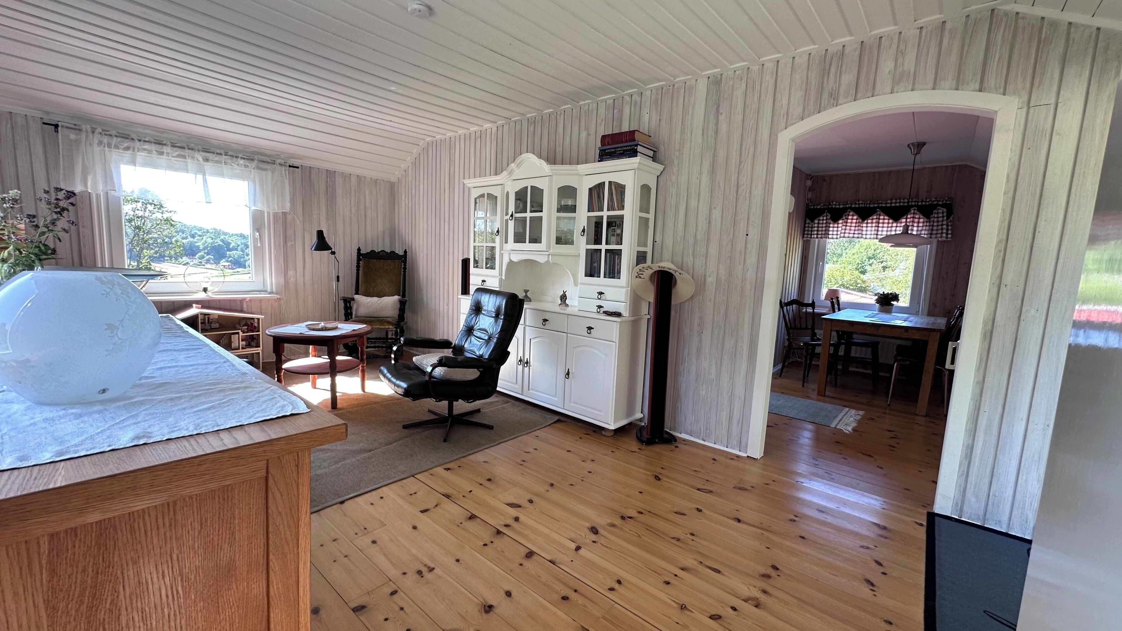 A room with a wooden floor, a dining area with a table and chairs, a white cabinet with glass doors, and a large window with curtains.