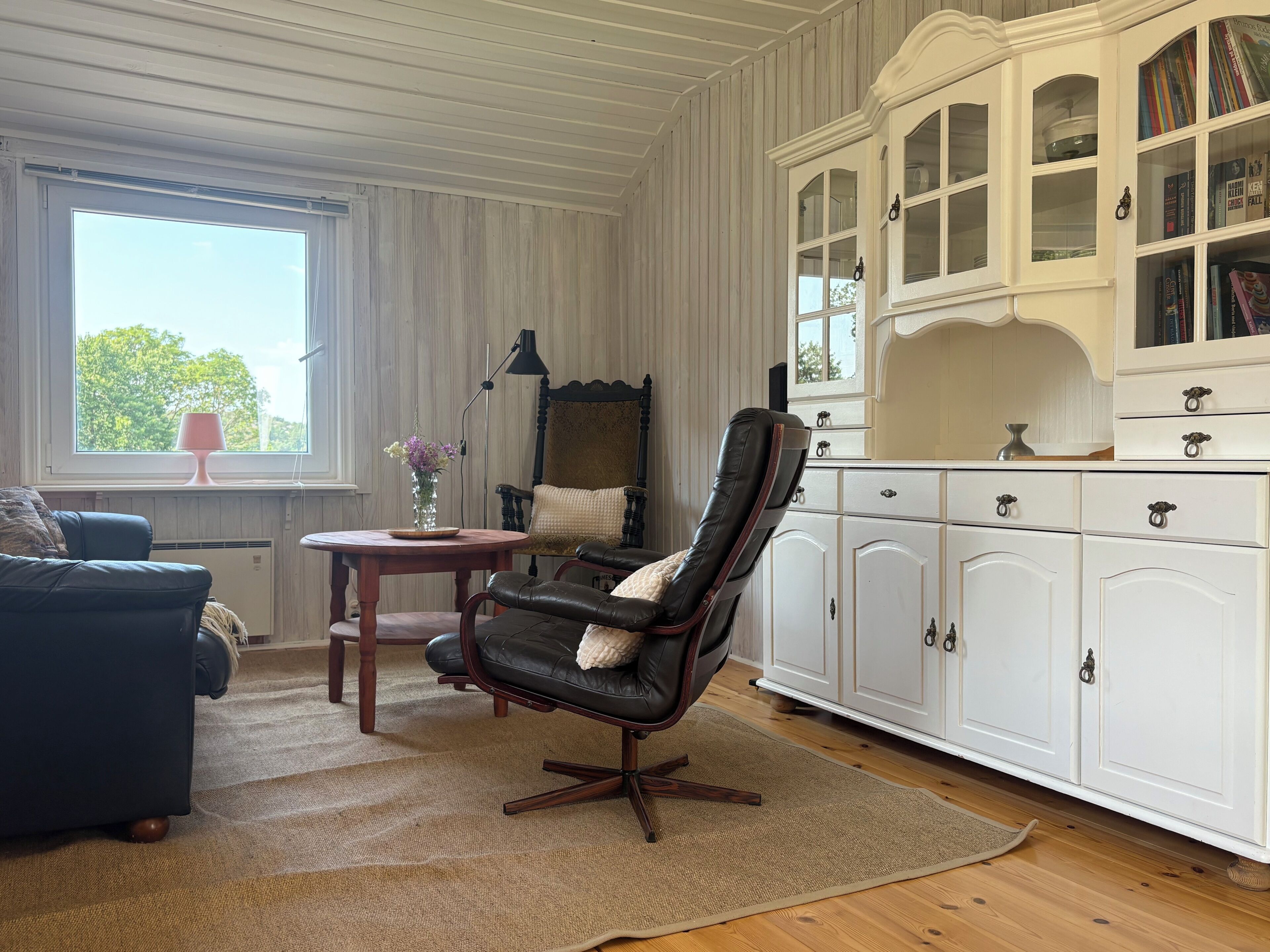 A room with a leather armchair, a wooden table, a white cabinet with glass doors, and a window with a view of trees.