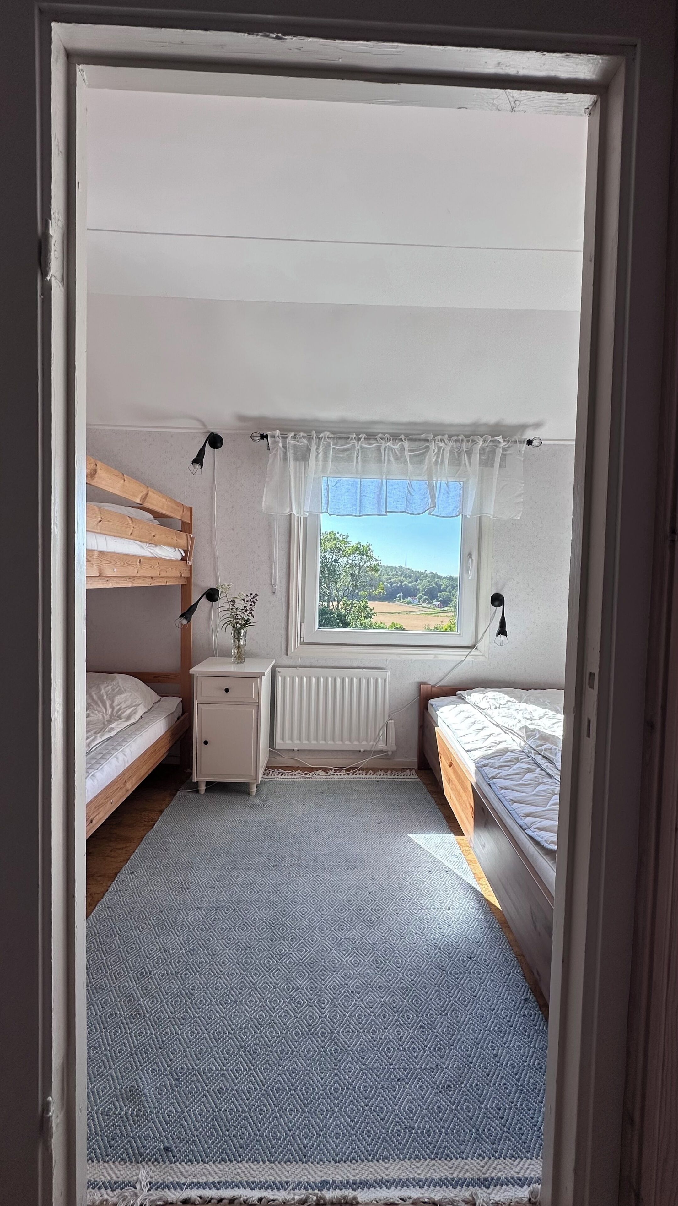 A bedroom with two beds, a radiator, a window with a view, and a bedside table with a lamp.