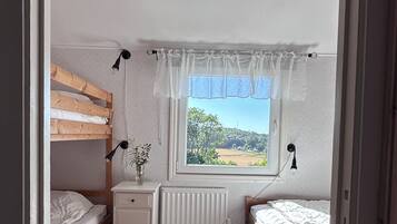 A bedroom with two beds, a radiator, a window with a view, and a bedside table with a lamp.