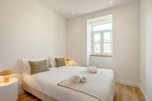 Ein Schlafzimmer mit einem Bett, Kissen, einer Nachttischlampe, einem Fenster und einem aufgerollten Handtuch auf dem Bett.