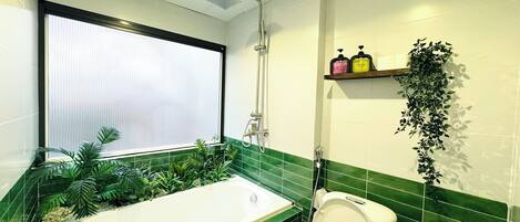 A bathroom with a bathtub, toilet, and shower. There is a window with blinds, a shelf with toiletries, and a plant.
