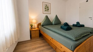 Un dormitorio con una cama de madera, una mesita de noche con lámpara, una planta en maceta y un cuadro en la pared.