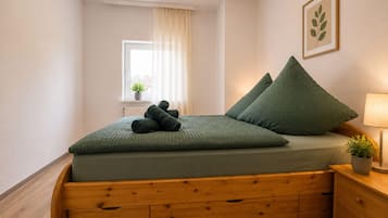 A bed with a wooden frame and a green mattress, a bedside table with a lamp, a window with curtains, and a framed plant on the wall.