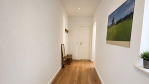 Un couloir avec un plancher en bois, des murs blancs, une porte au fond et un tableau encadré accroché au mur.