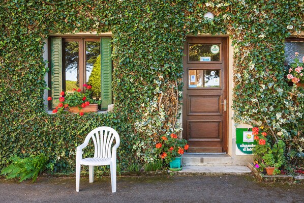 A building with a wooden door and a window with shutters, surrounded by lush greenery and potted plants.