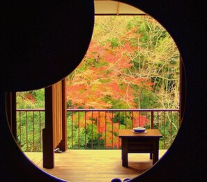 A circular window view of a balcony with a table and a chair, overlooking a garden with trees. - Hakone Shiunso (Hakone)