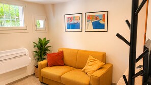 Ein Zimmer mit einem gelben Sofa, einer Topfpflanze und zwei gerahmten Kunstwerken an der Wand.