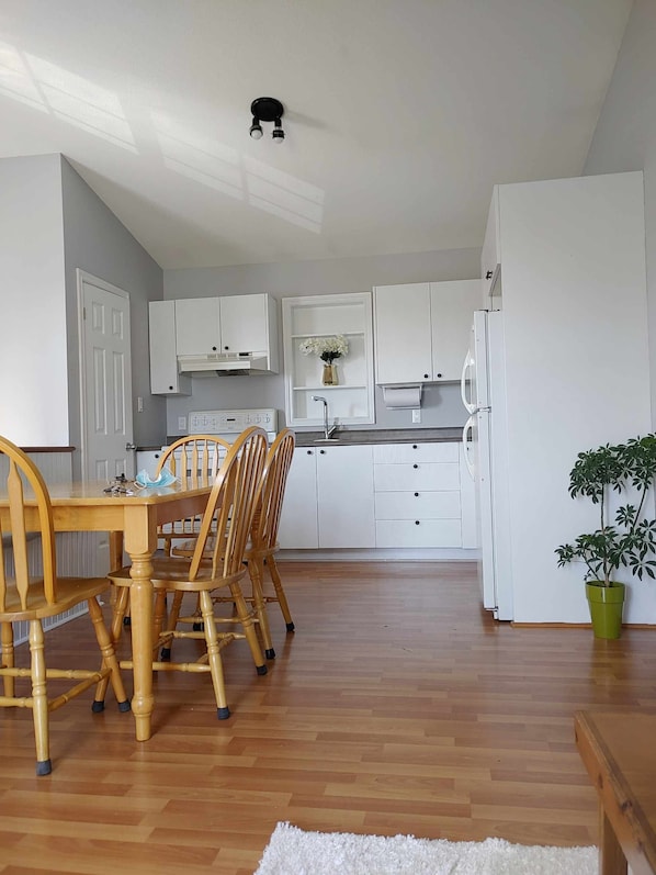 A kitchen with white cabinets, a wooden dining table with chairs, a refrigerator, and a potted plant.
