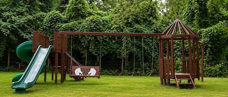 Une aire de jeux avec un toboggan, des balançoires et une cabane en bois.
