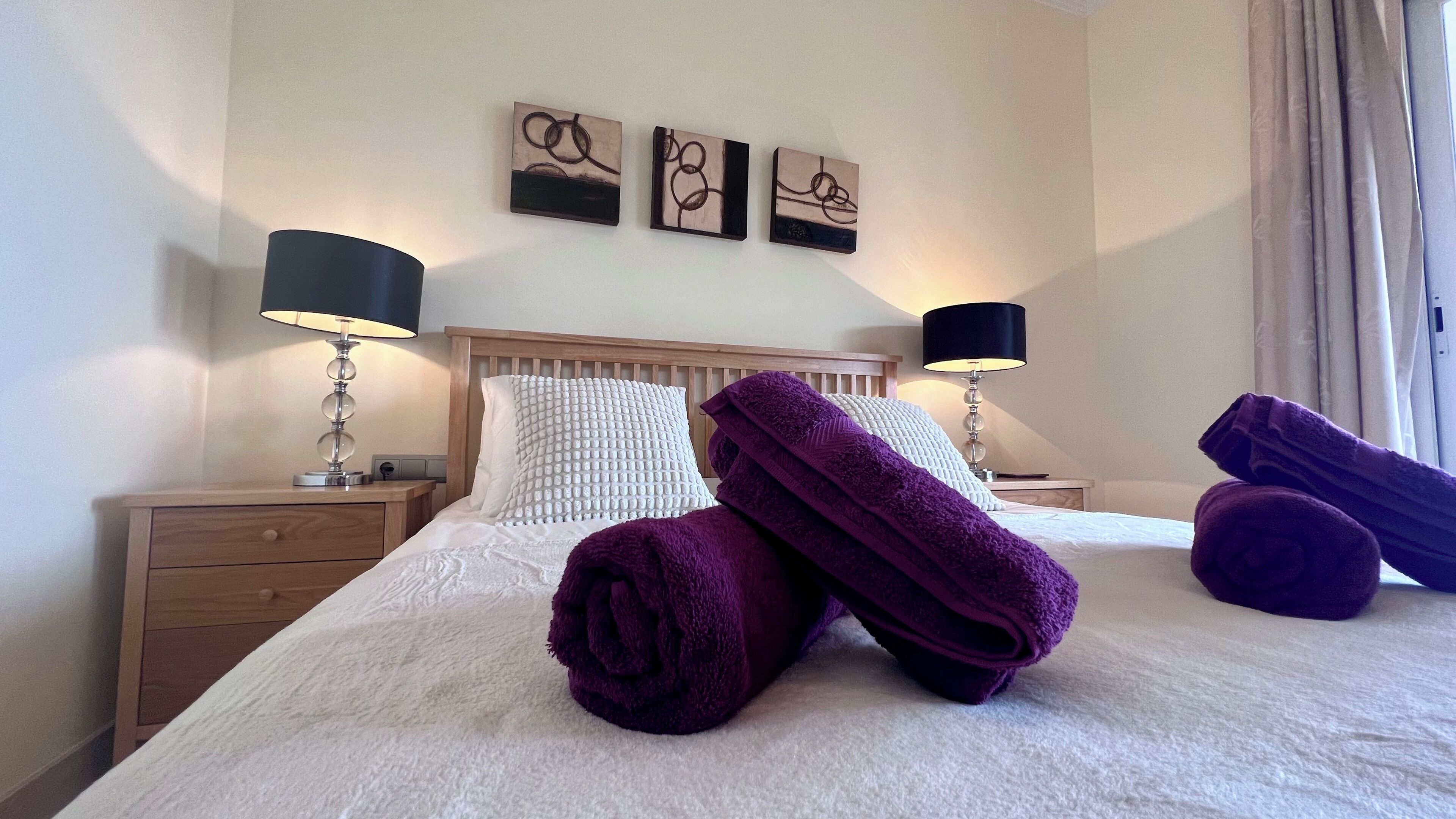 Habitación de hotel con cama, dos mesitas de noche, dos lámparas, cabecera y tres obras de arte en la pared.