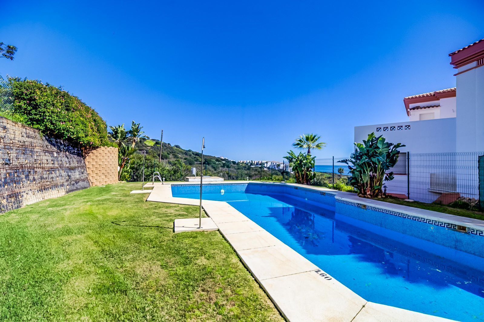 Una piscina con vista despejada al océano, rodeada por un césped cuidado y un muro de piedra.