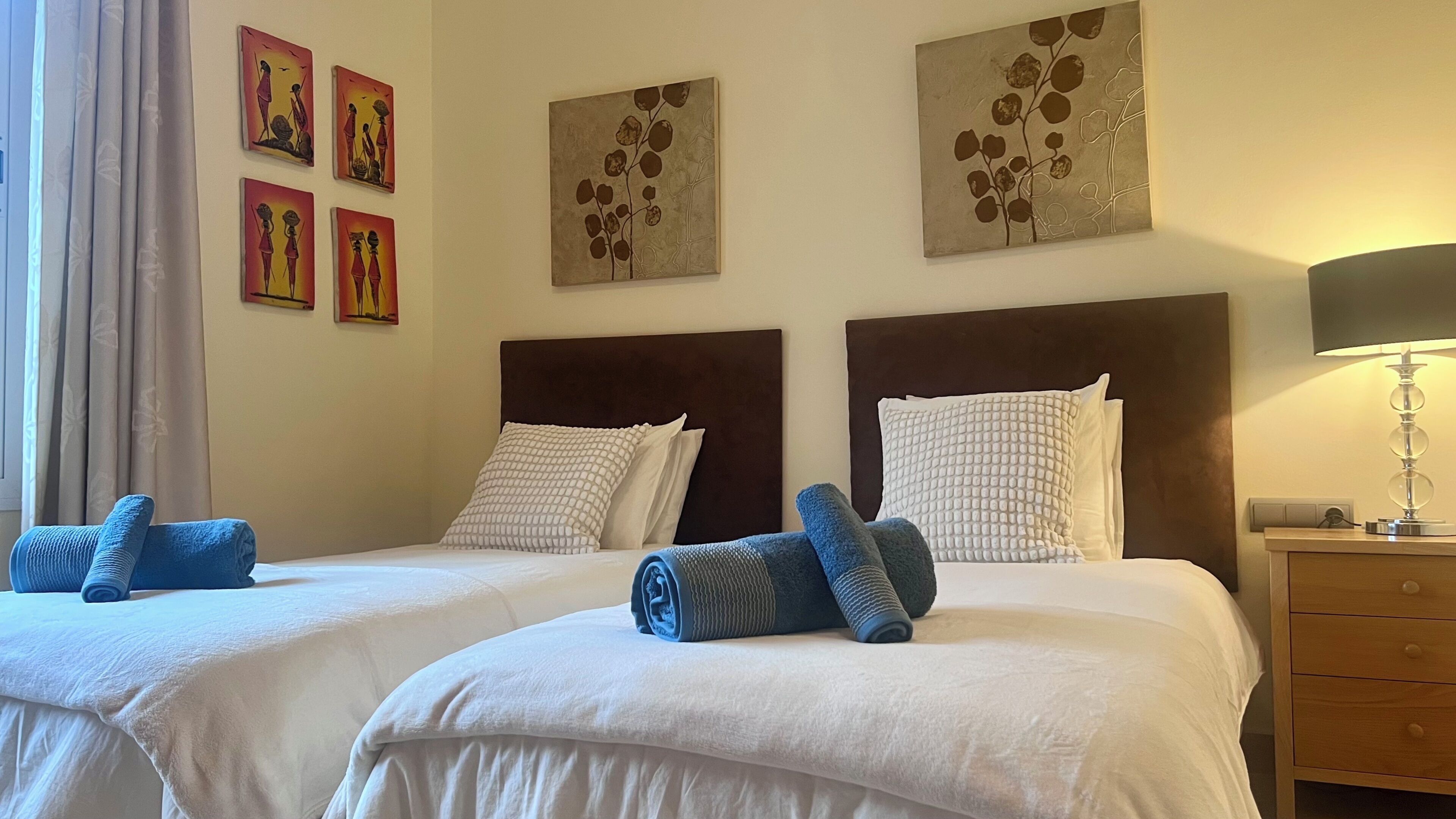 A hotel room with two beds, each with a headboard, white bedding, and blue towels. There are wall art pieces above the beds and a lamp on the nightstand.