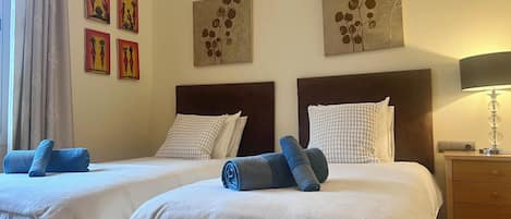A hotel room with two beds, each with a headboard, white bedding, and blue towels. There are wall art pieces above the beds and a lamp on the nightstand.