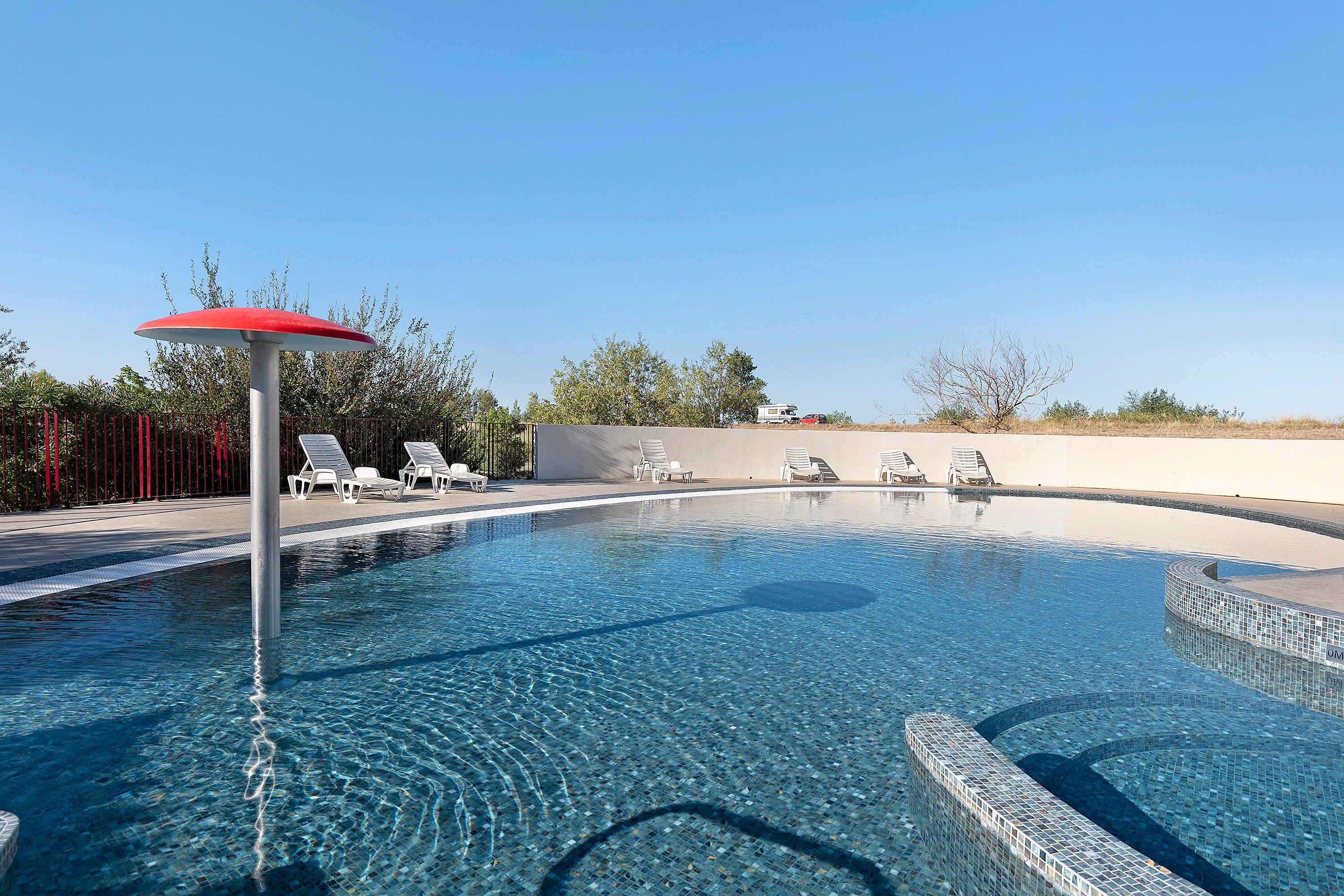 A swimming pool with clear water, surrounded by lounge chairs and a red umbrella.