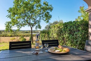 An outdoor table set with a bottle of wine, glasses, cheese, grapes, and bread, with a view of a tree and landscape in the background. - Contemporary house with lake view near Lake Jouarres (Homps)