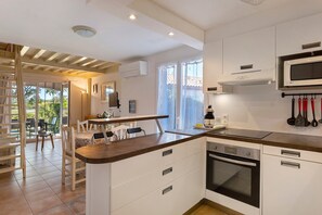 A modern kitchen with white cabinetry, a built-in oven, and a microwave. There is a dining area with a table and chairs, and a view of the outdoors through large windows. - Contemporary house with lake view near Lake Jouarres (Homps)