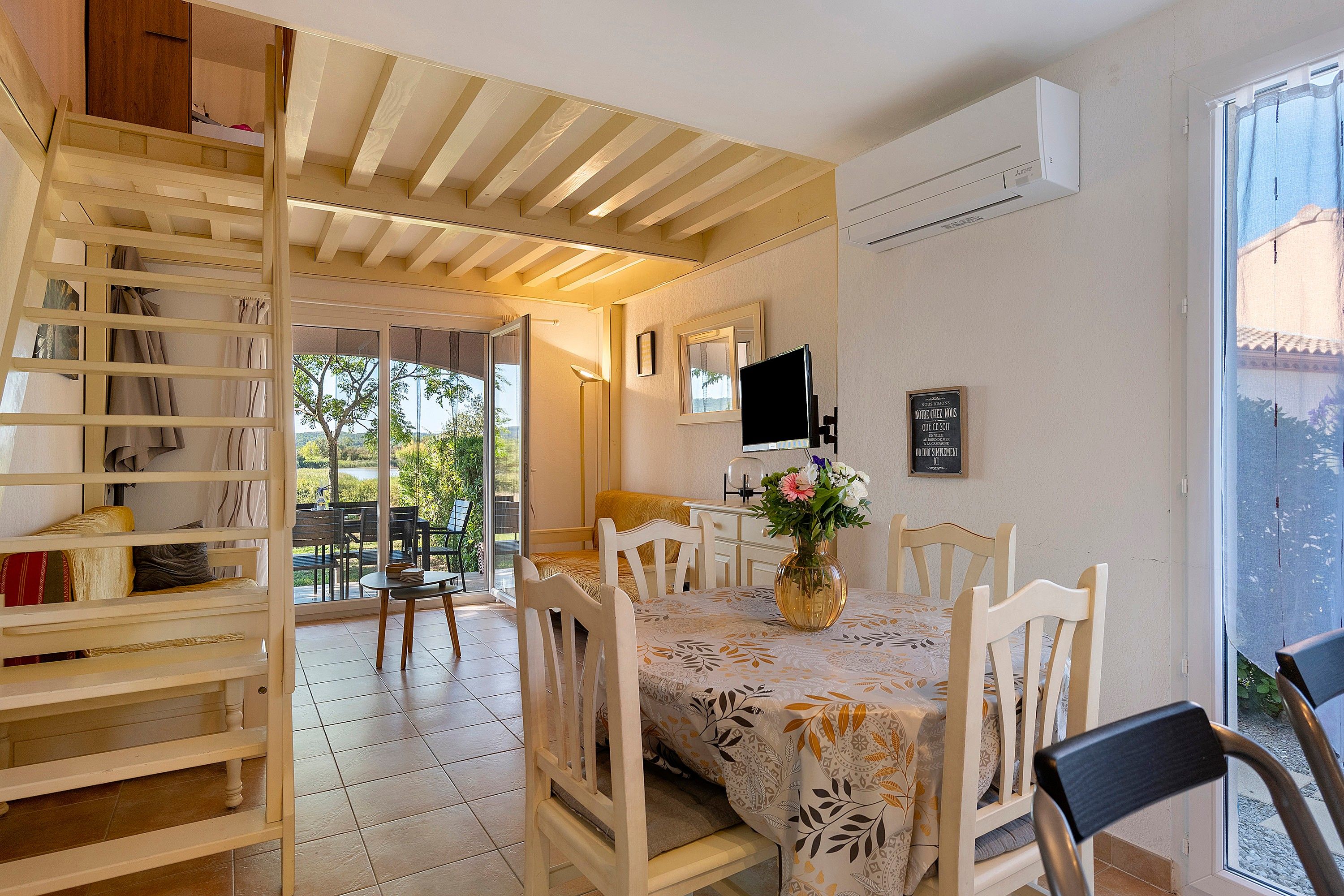 Un comedor con una mesa lista para cuatro, un televisor montado en la pared y una ventana con vista a follaje.