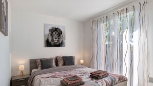 A bedroom with a bed, pillows, a nightstand, a lamp, a wall-mounted picture, and sheer curtains.