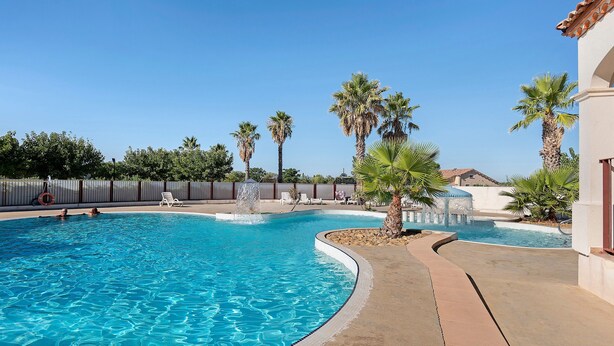 A swimming pool with clear blue water, surrounded by palm trees and a tiled deck.
