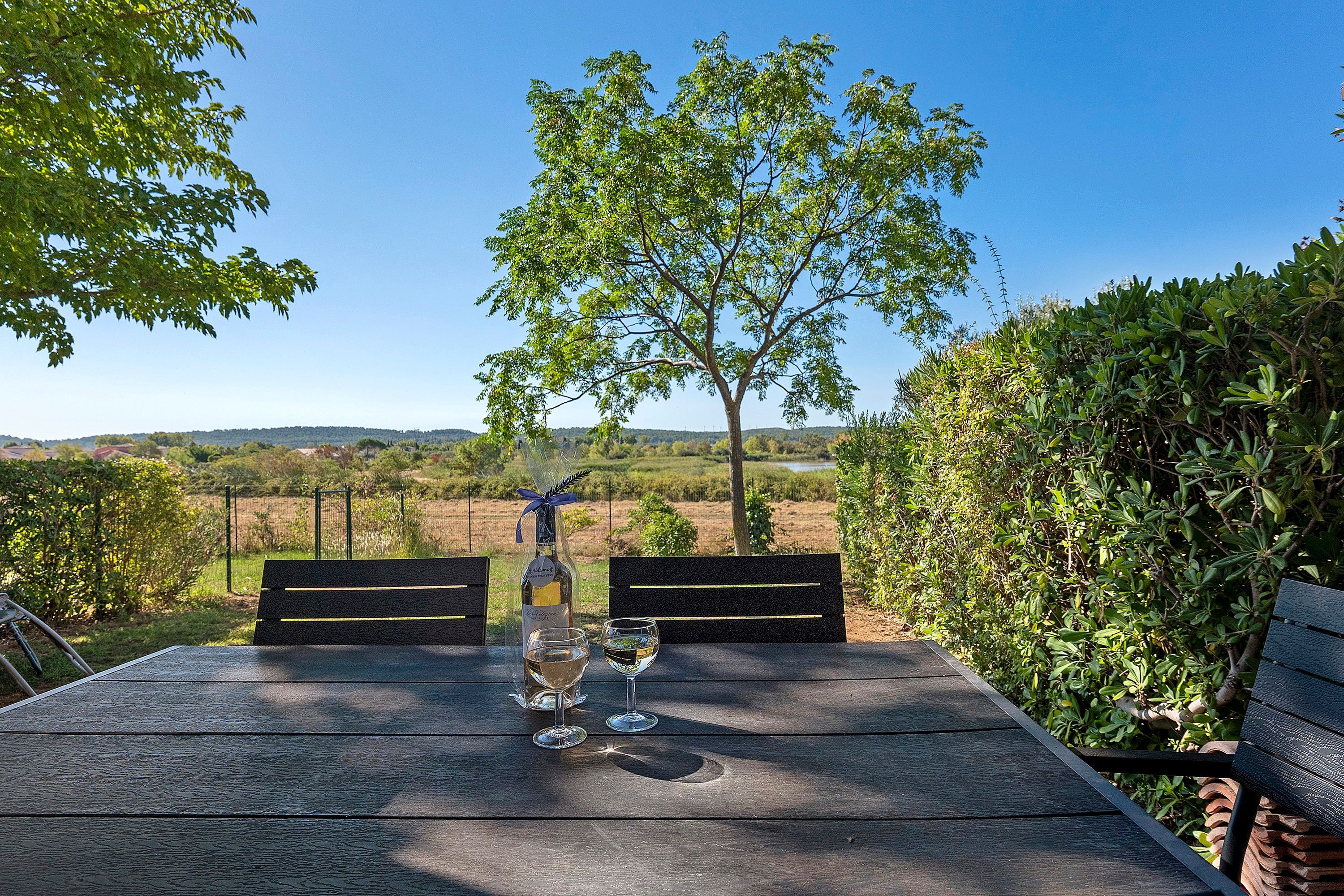 Une table en bois, deux chaises, une bouteille de vin et deux verres, le tout installé à l’extérieur, avec vue sur un arbre et un paysage.