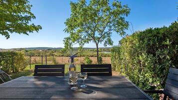 Une table en bois, deux chaises, une bouteille de vin et deux verres, le tout installé à l’extérieur, avec vue sur un arbre et un paysage.