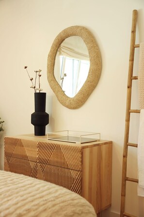 Un dormitorio con un aparador de madera, un espejo redondo, un jarrón negro con flores secas y una escalera de madera.